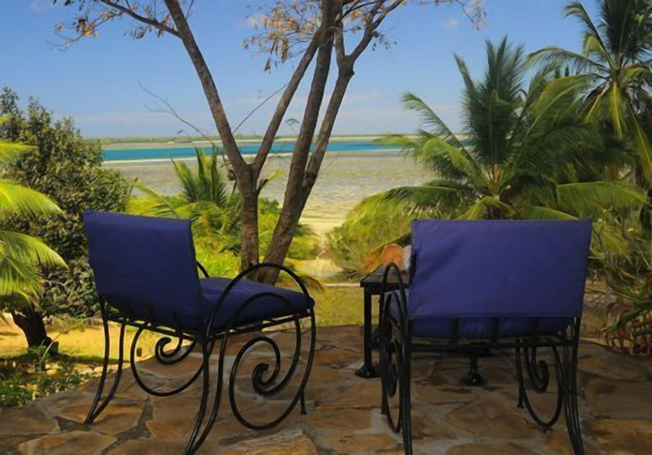 Deux chaises bleues à armature métallique, placées sur une terrasse en pierre donnant sur une plage bordée de palmiers et surplombant un ciel dégagé.