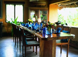 Een eettafel is gedekt met blauw glaswerk en een vaas met bloemen, omringd door stoelen.