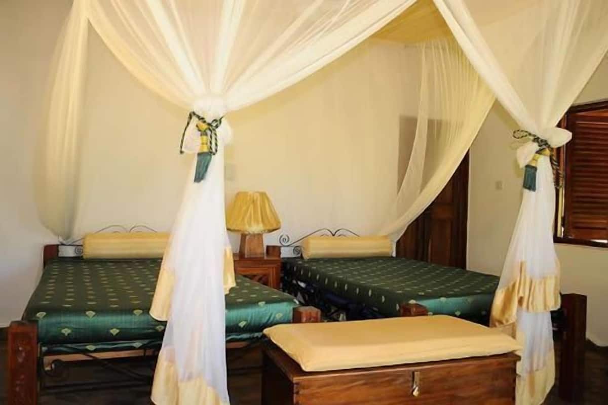 A room with two single beds, each with a green bedspread and a wooden headboard. There is a wooden nightstand with a lamp between the beds. The room has a white canopy bed with a sheer white curtain.