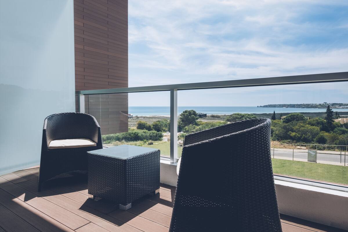 A balcony with wicker furniture, a glass railing, and a view of the sea and greenery.