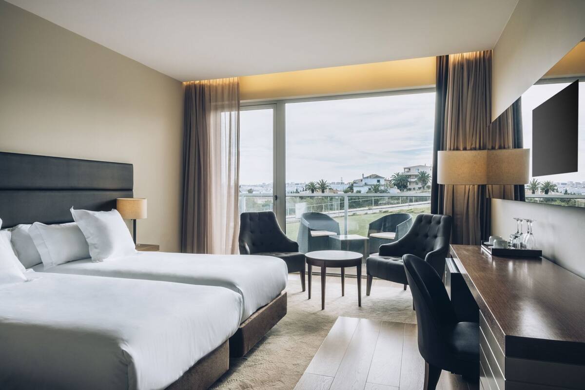 A hotel room with a large bed, a desk, two chairs, a TV, and a balcony with a city view.