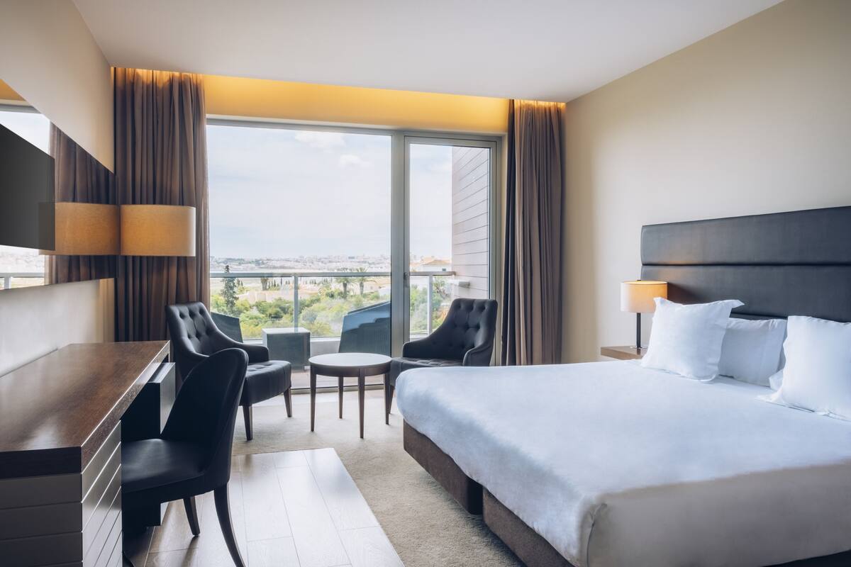 A hotel room with a bed, a desk, two chairs, a small table, and a view of the city through large windows.
