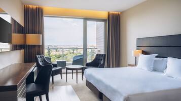 Habitación de hotel con una cama, un escritorio, dos sillas, una mesita y vistas a la ciudad a través de amplios ventanales.