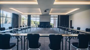 Un salón de conferencias con mesas largas y sillas, ventanas amplias y una pantalla para proyector.