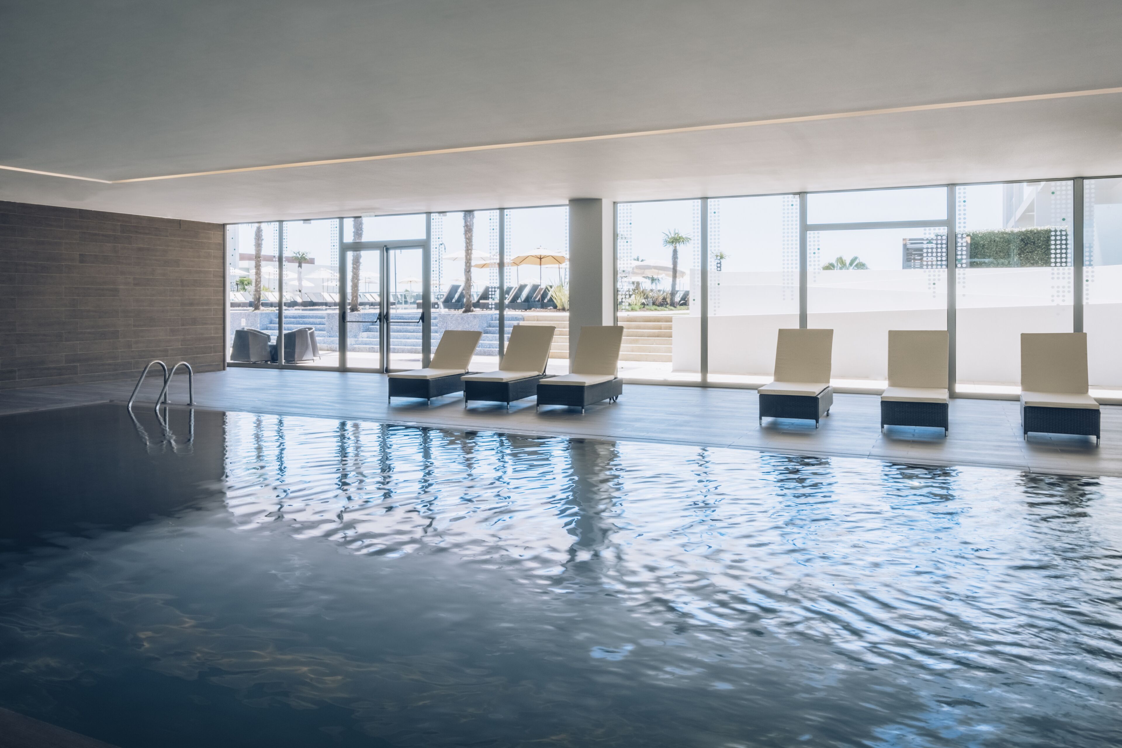 An indoor swimming pool with clear glass walls, lounge chairs, and a brick wall.