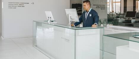 Un mostrador de recepción con una mampara de vidrio, un hombre de pie detrás y un cartel que enumera servicios como 'Quartos Pares / Even Rooms' y 'Bar Piscina / Pool Bar'.