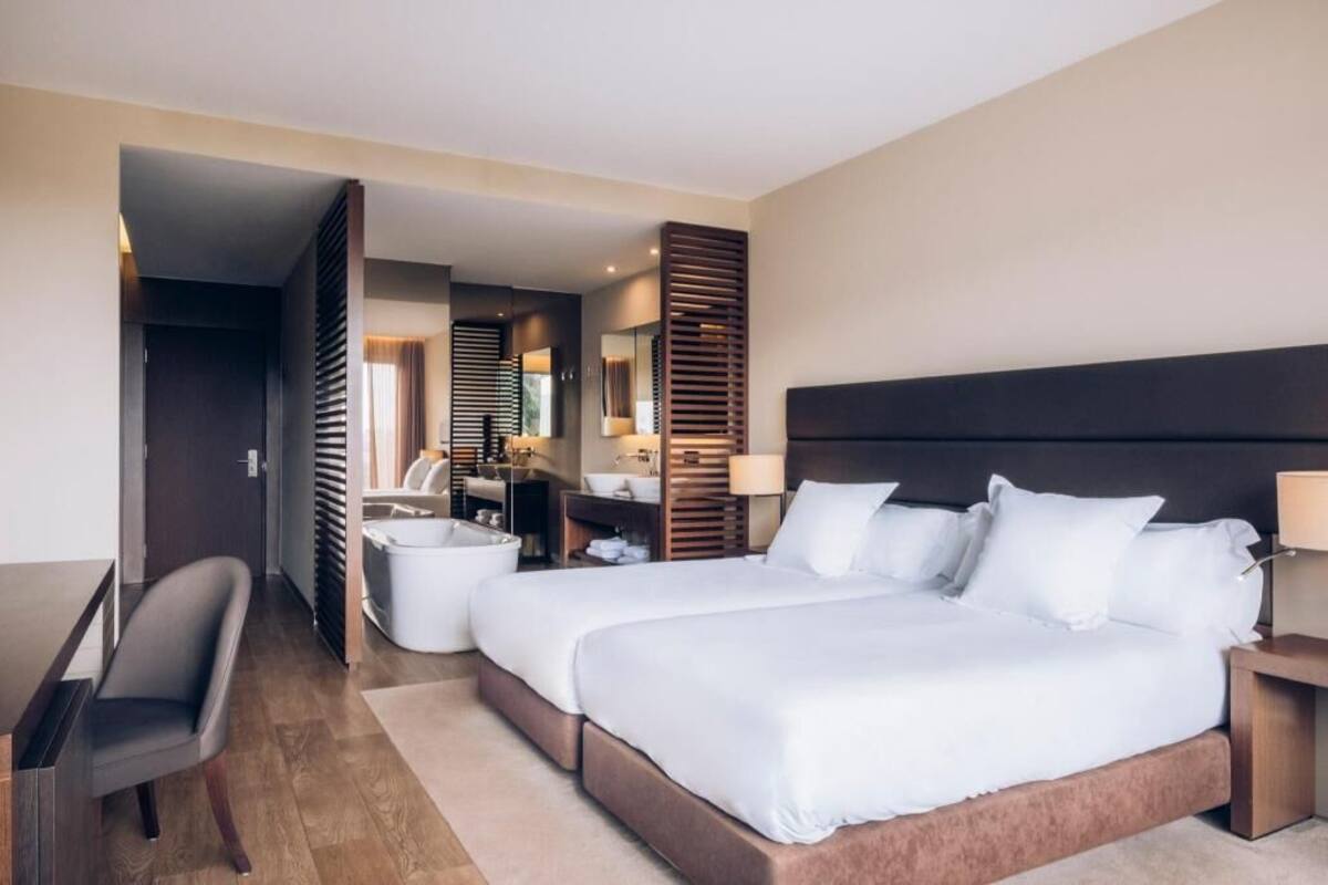 A hotel room with a large bed, a desk, a chair, a bathroom with a sink and mirror, and a view of the outside.