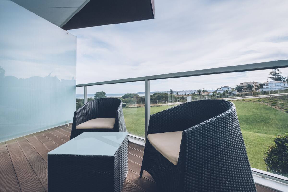 A balcony with wicker chairs and a table, overlooking a grassy area and buildings.
