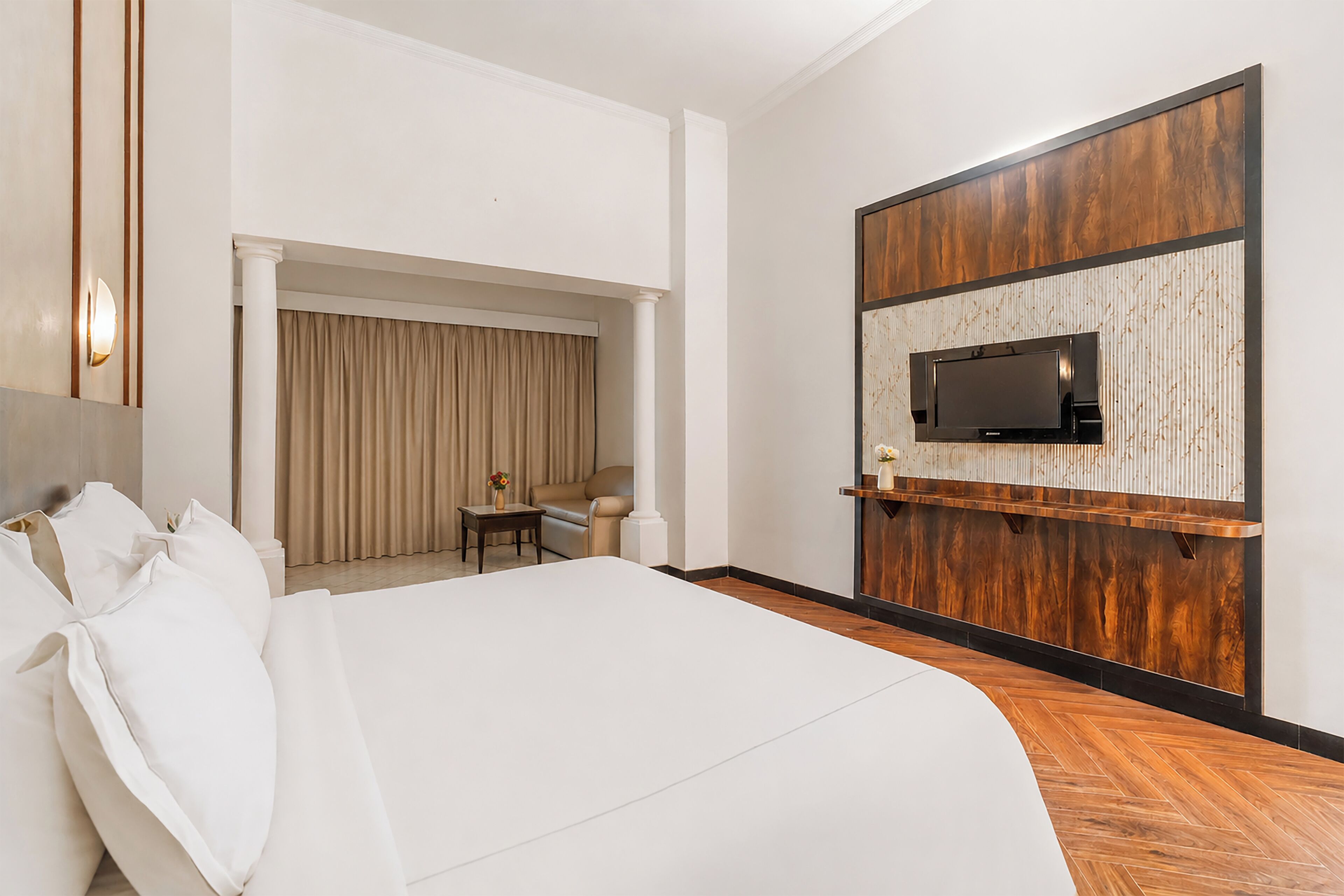 A hotel room with a large bed, a TV mounted on a wooden panel wall, a small table with a chair, and a window with curtains.