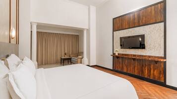 A hotel room with a large bed, a TV mounted on a wooden panel wall, a small table with a chair, and a window with curtains.