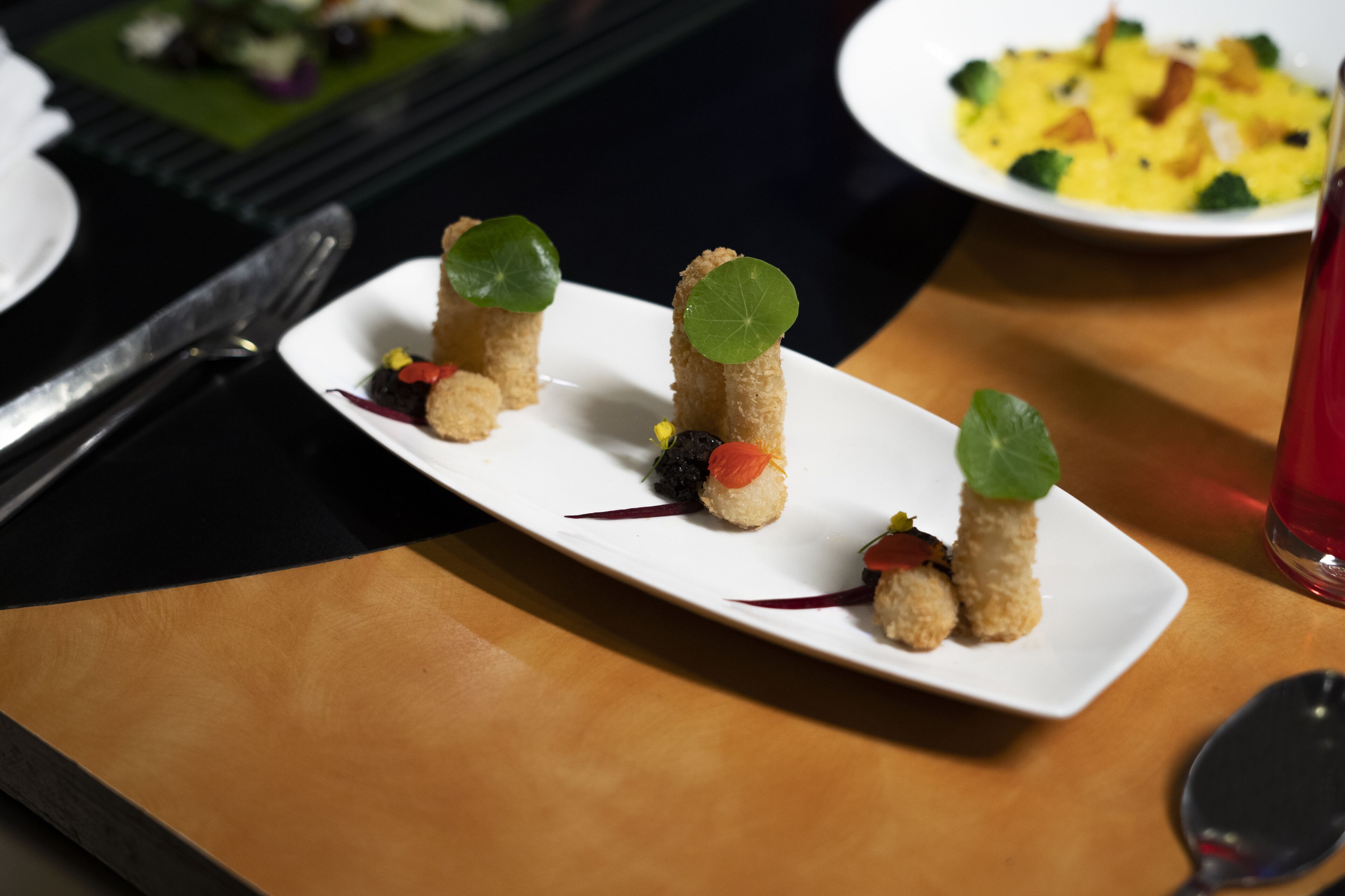 A white rectangular plate with three small, cylindrical food items, each topped with a leaf, on a wooden table.