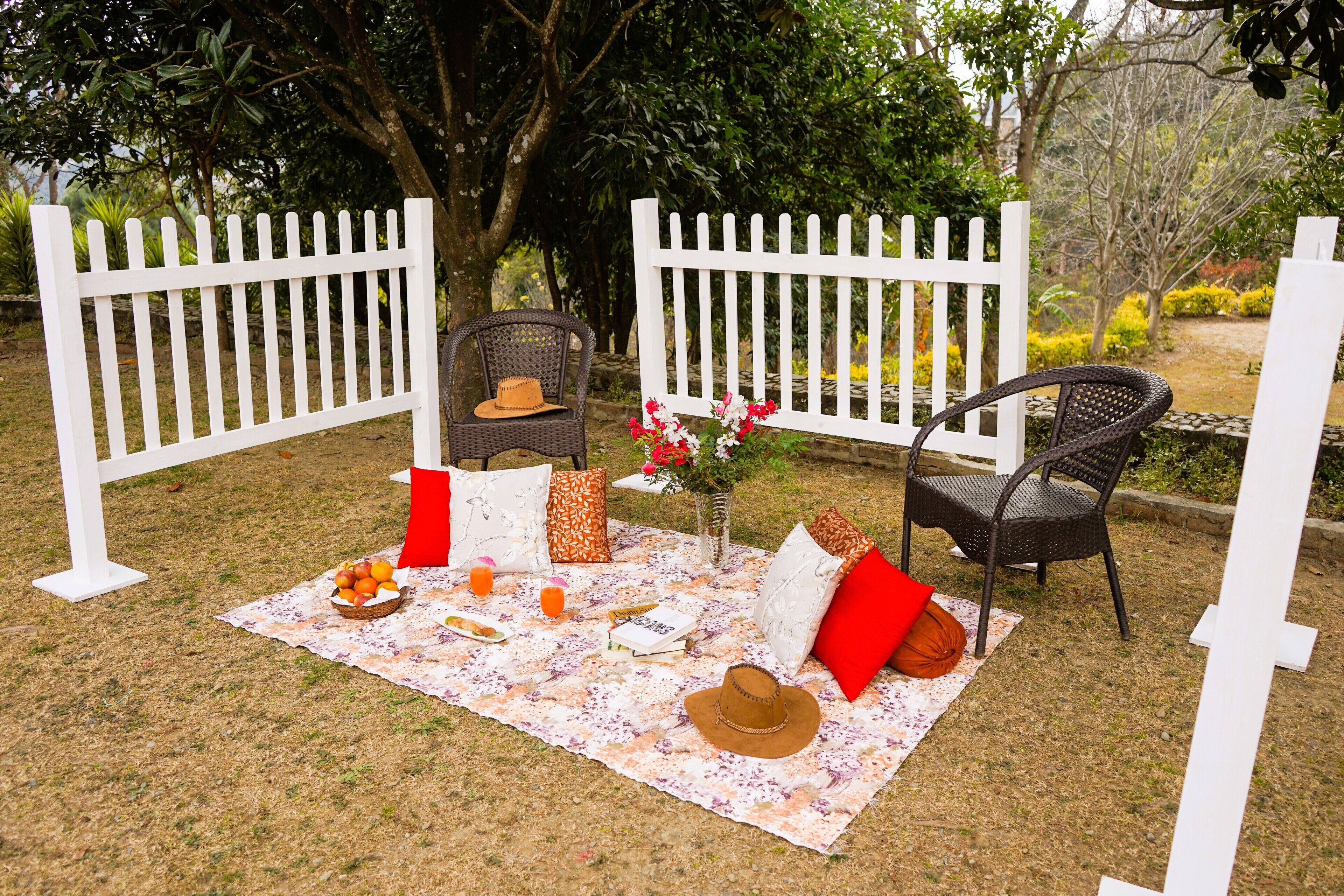 A picnic setup with a blanket, chairs, a hat, a bowl of fruit, and a vase of flowers.