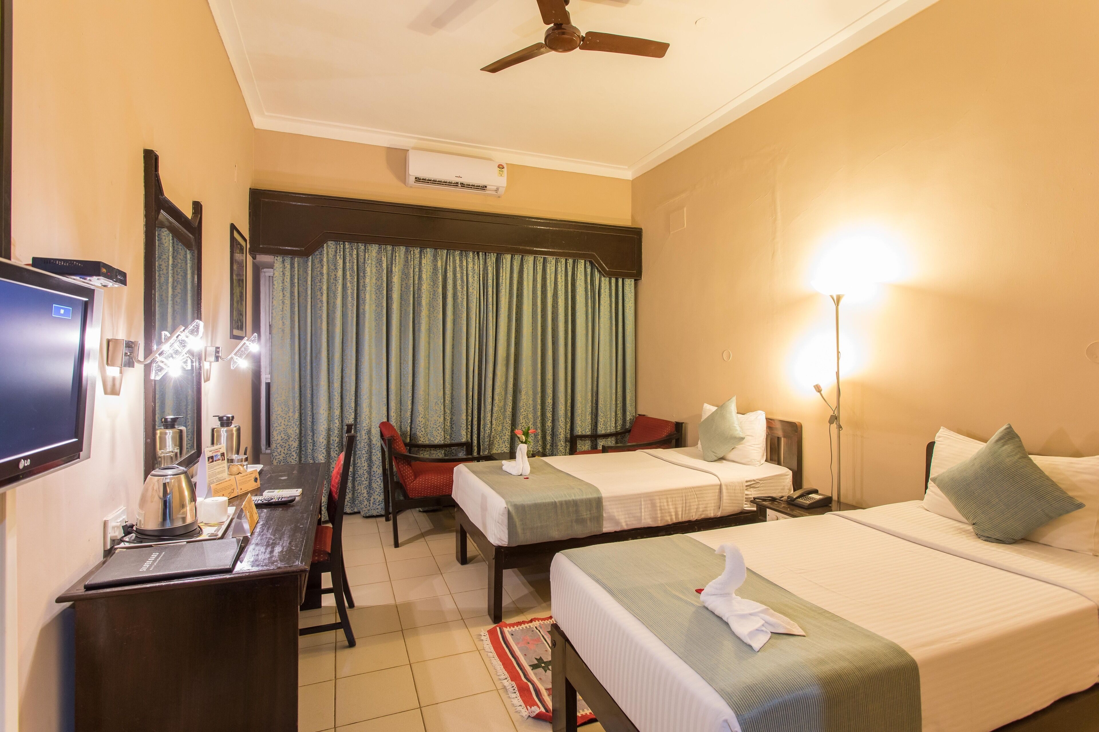 A hotel room with two beds, a desk with a television, a ceiling fan, and a window with curtains.