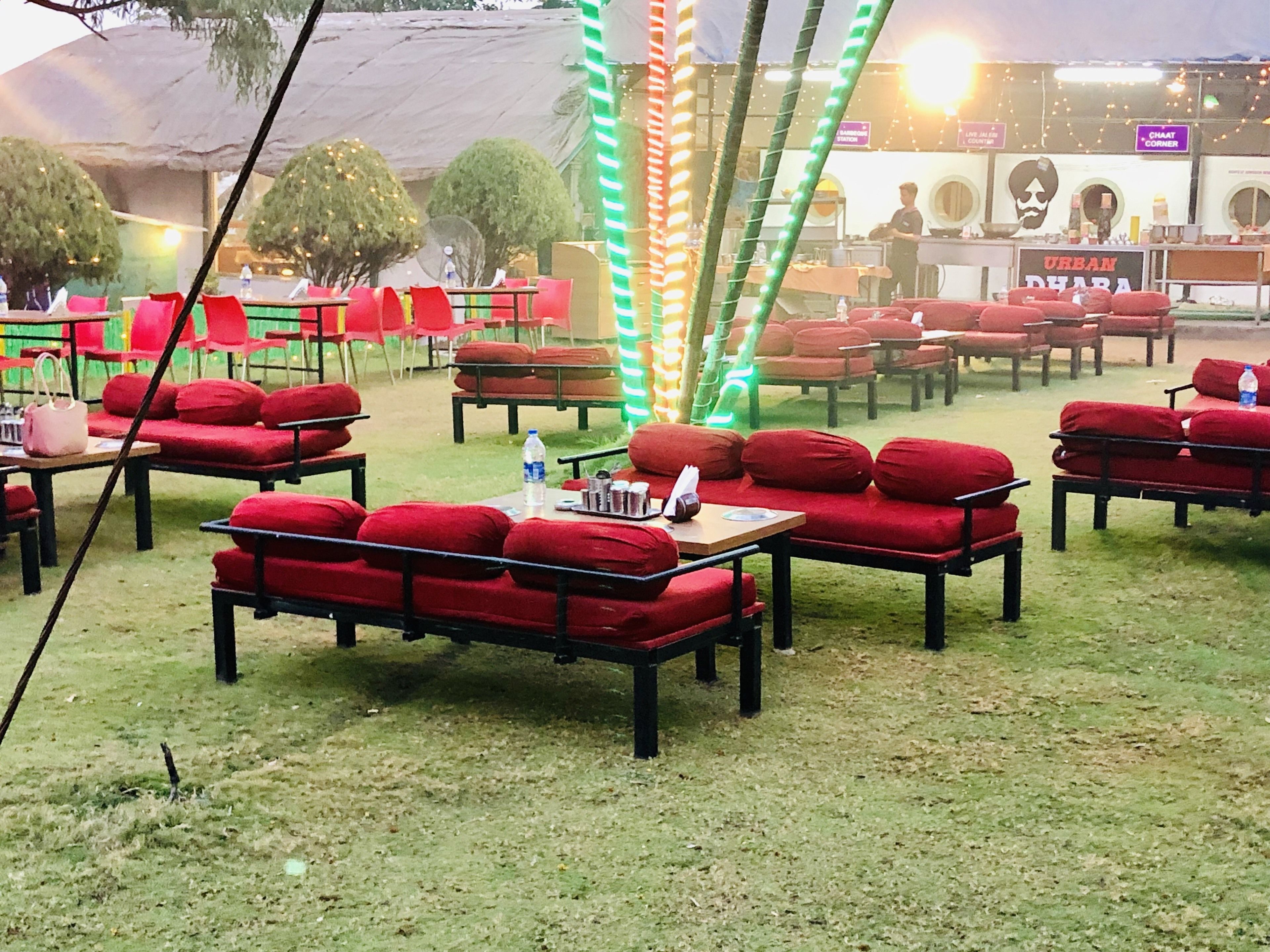 Un área de estar al aire libre con bancos rojos acolchados y mesas, rodeada de árboles y luces colgantes.