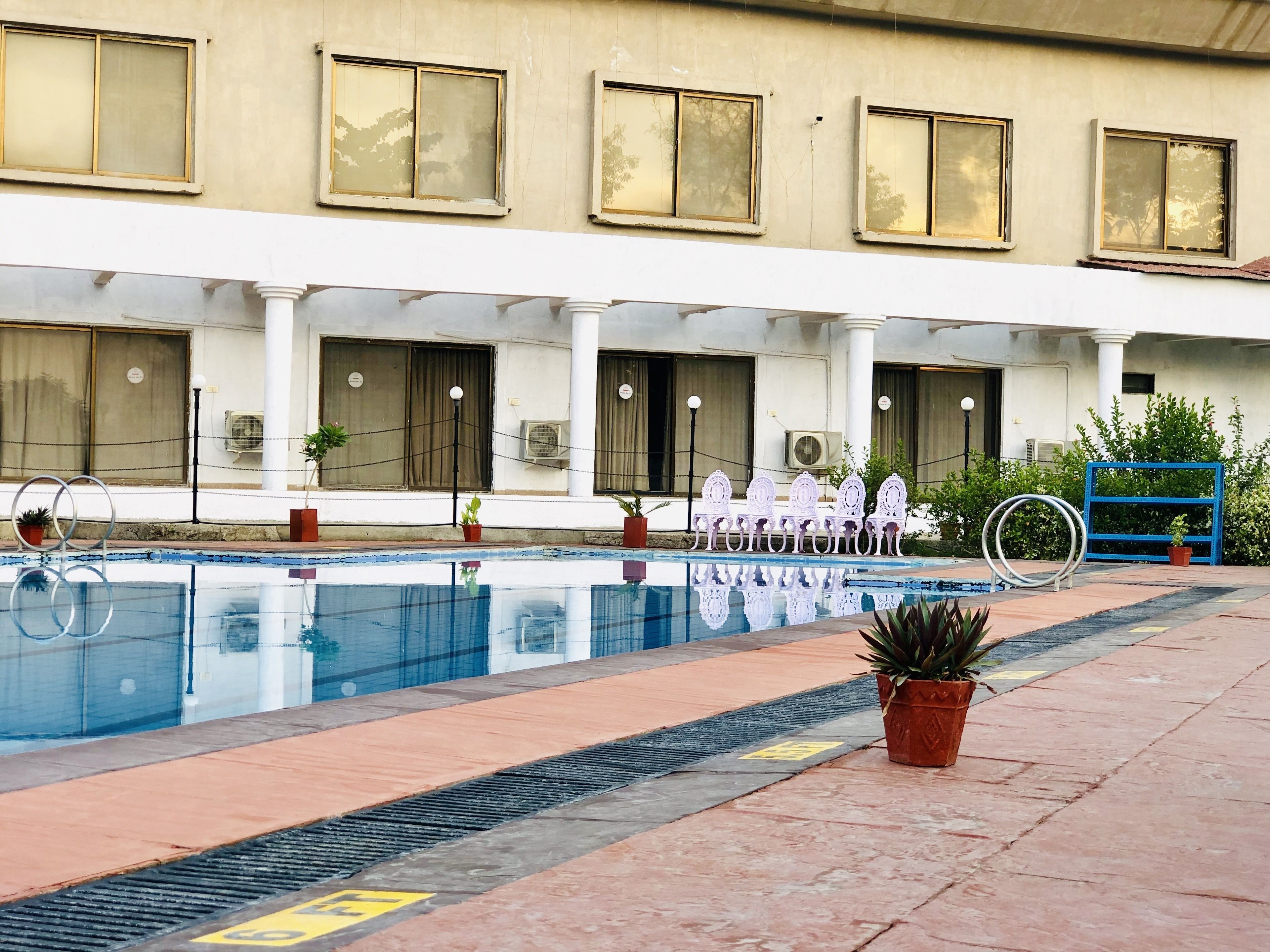 Un espace piscine offrant une vue dégagée sur la piscine, un coin salon avec des chaises, et un bâtiment doté de nombreuses fenêtres et de plusieurs unités de climatisation.