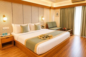 Una habitación de hotel con una cama grande, una mesita de noche con lámpara, una silla, una pequeña mesa con fruta y una ventana con cortinas.