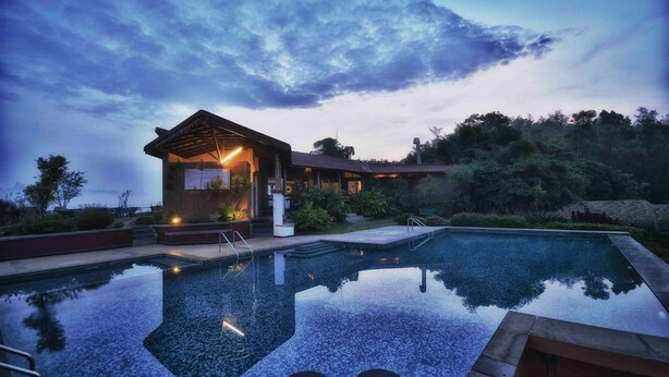 A swimming pool with a wooden deck and a building with a sloped roof in the background. - Indo Asia Heritage, Coorg (Madikeri)