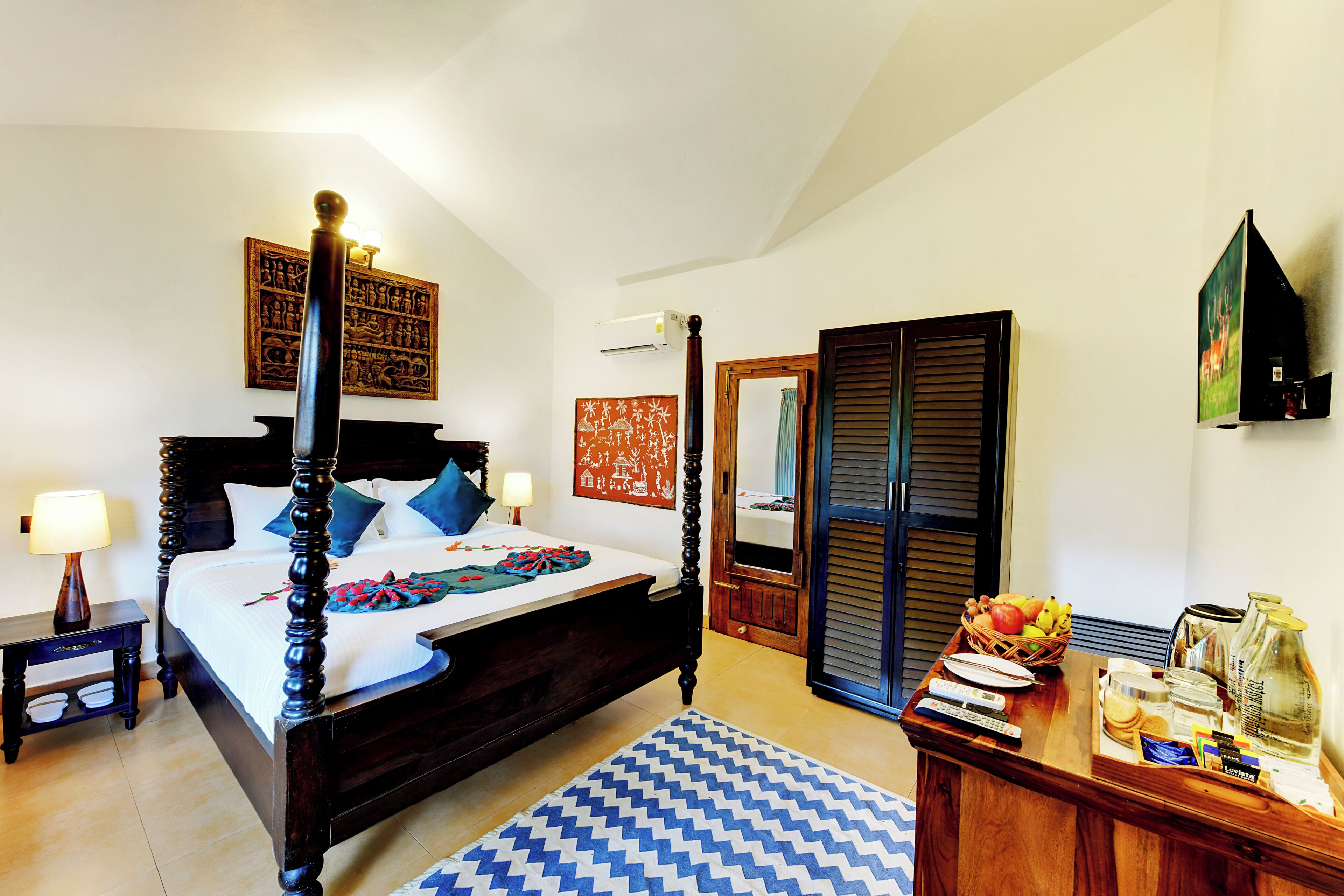 A four-poster bed with a blue and white rug, a wooden nightstand, a bedside table with a lamp, a wooden wardrobe, and a breakfast tray with fruits and a bottle.