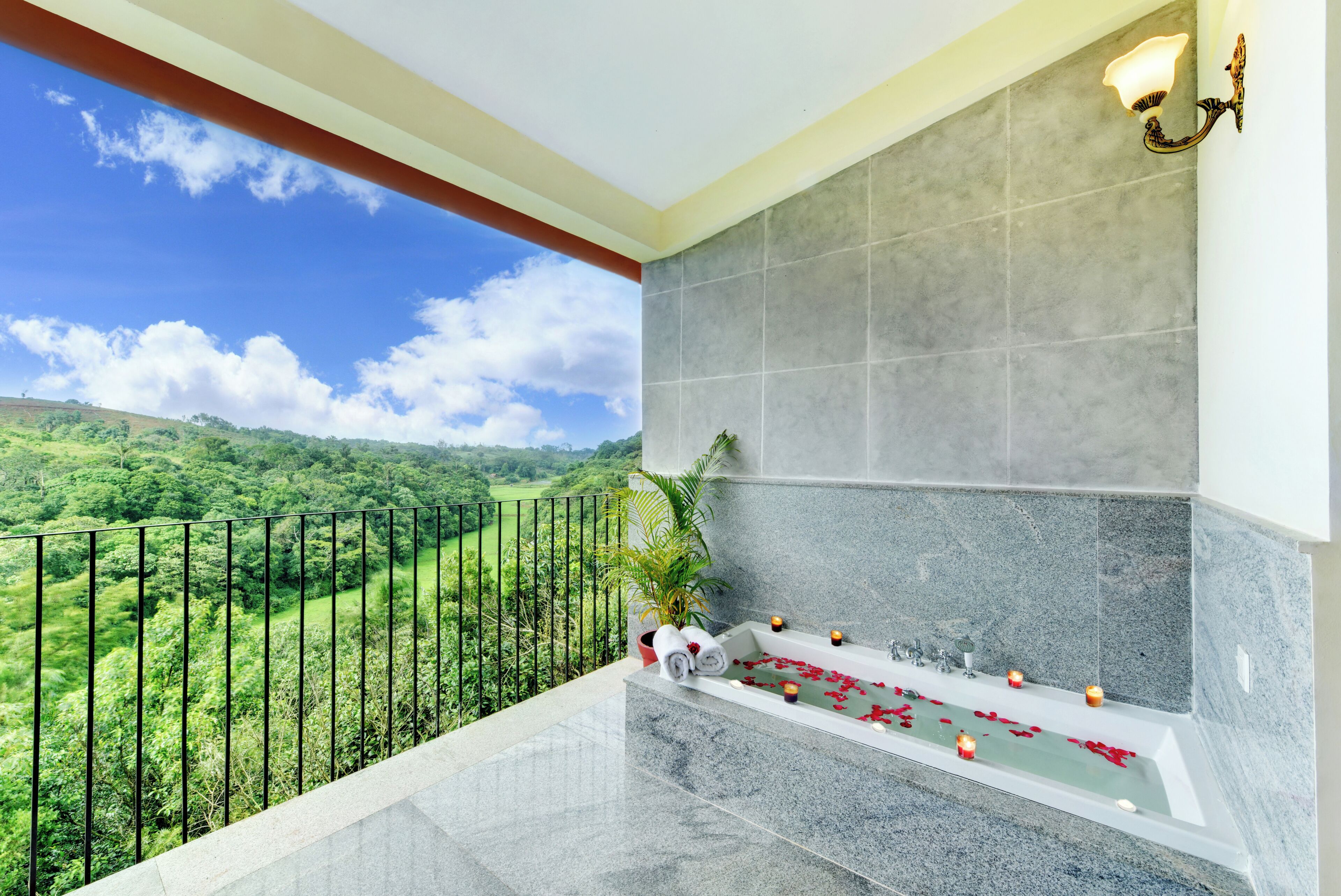 A balcony with a view of a green landscape, a bathtub with rose petals, and a wall-mounted light fixture.