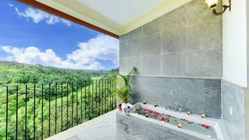 A balcony with a view of a green landscape, a bathtub with rose petals, and a wall-mounted light fixture.