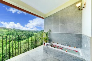 Un balcón con vista a un paisaje verde, una bañera con pétalos de rosa y una lámpara fijada a la pared.