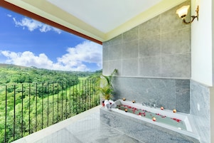 Un balcone con vista su un paesaggio verde, una vasca da bagno con petali di rosa e un lampadario a parete.
