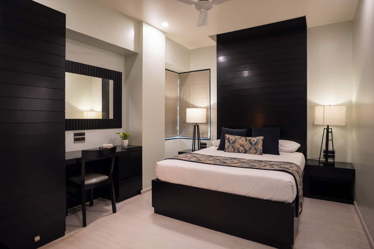 Une chambre moderne avec un grand lit, un bureau et une chaise. Il y a un miroir au mur et un ventilateur de plafond.