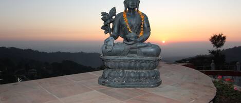 A statue of a seated figure with a flower, placed on a tiled platform with a sunset in the background.