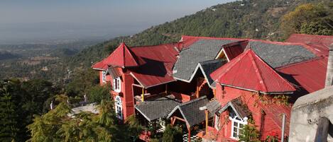 A red-roofed building with multiple levels and a balcony, surrounded by lush greenery and hills in the background.