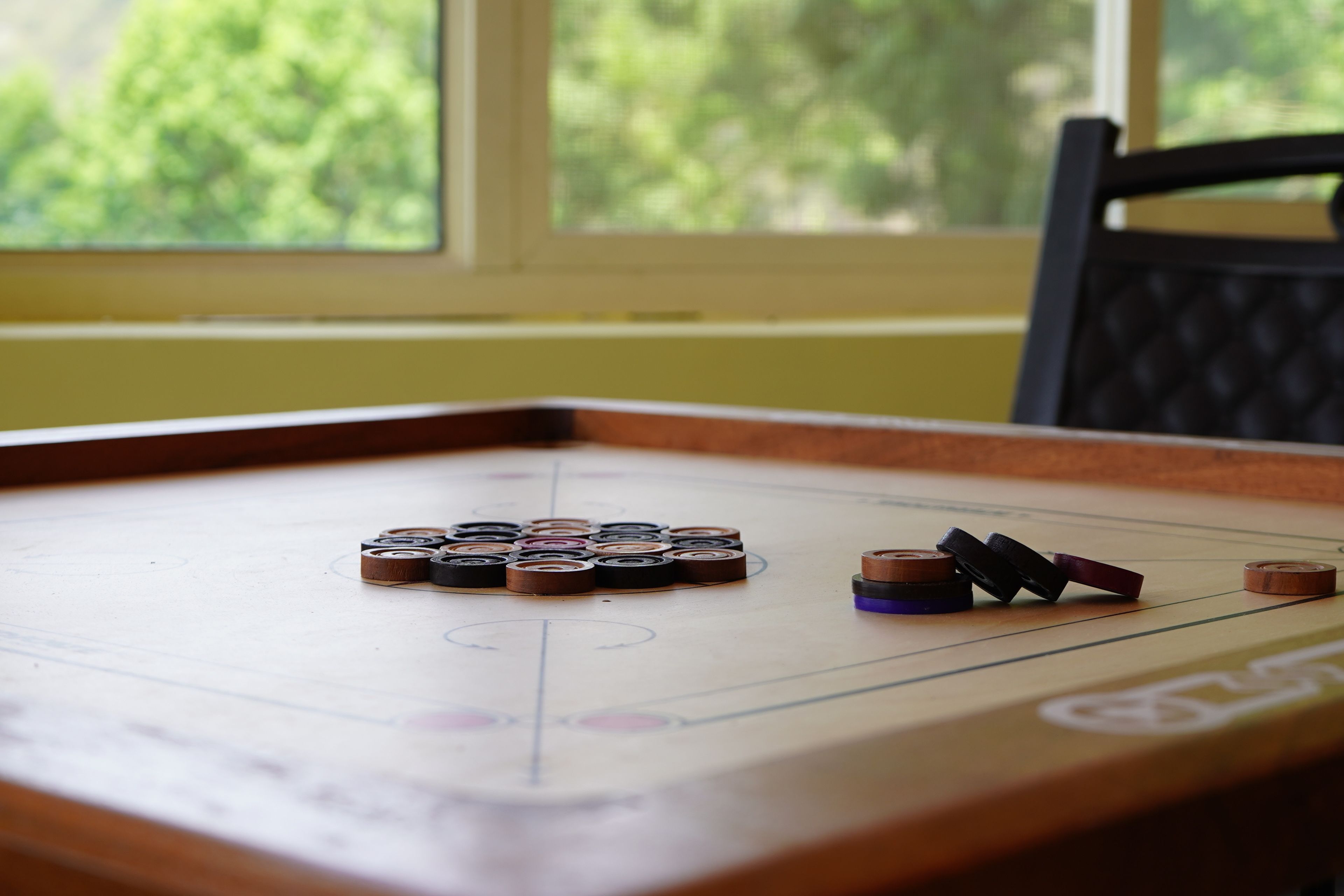 A shuffleboard table with pucks in various colors, positioned near a window with a view of greenery.