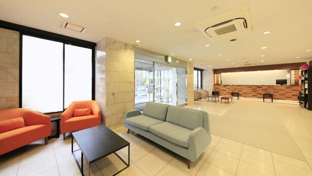 A spacious lobby with a light blue sofa, an orange armchair, and a black coffee table. There are large windows, a reception desk, and a fire extinguisher visible. - Smile Hotel Tokyo Ayase Ekimae (Tokyo)