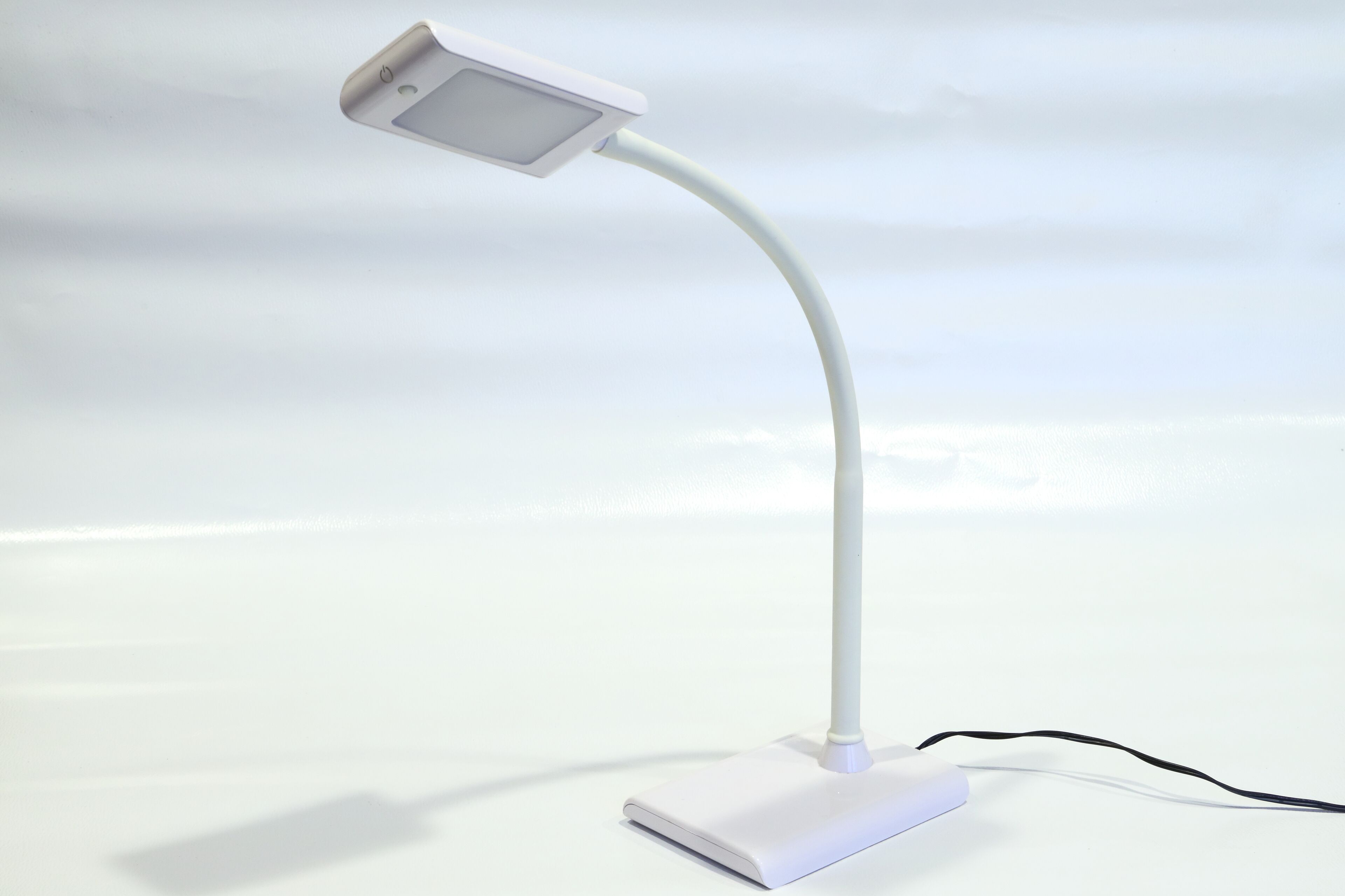 A white desk lamp with a curved arm and a rectangular base on a white surface.