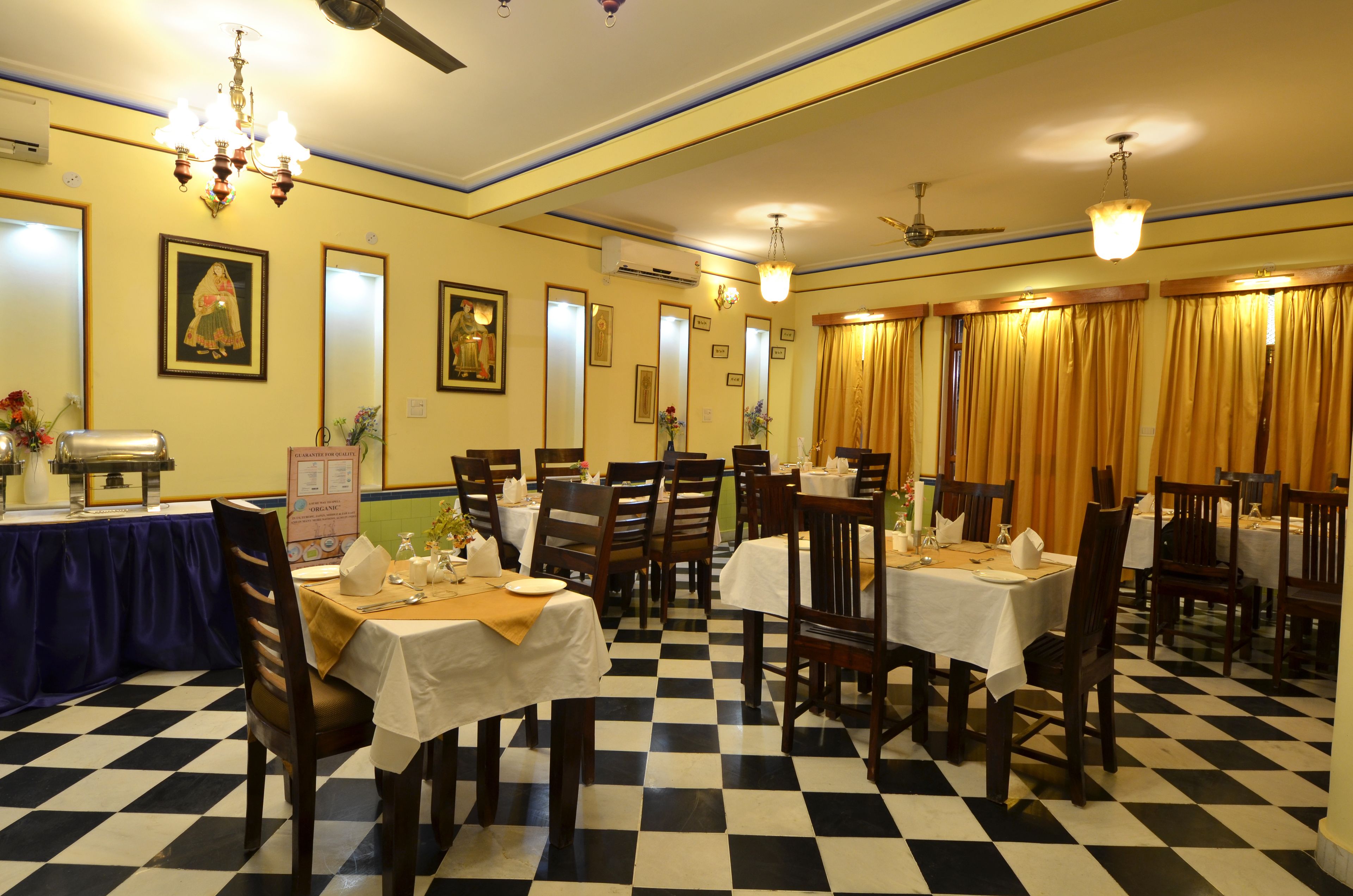 A dining area with checkered flooring, tables with white tablecloths, wooden chairs, and a chandelier.