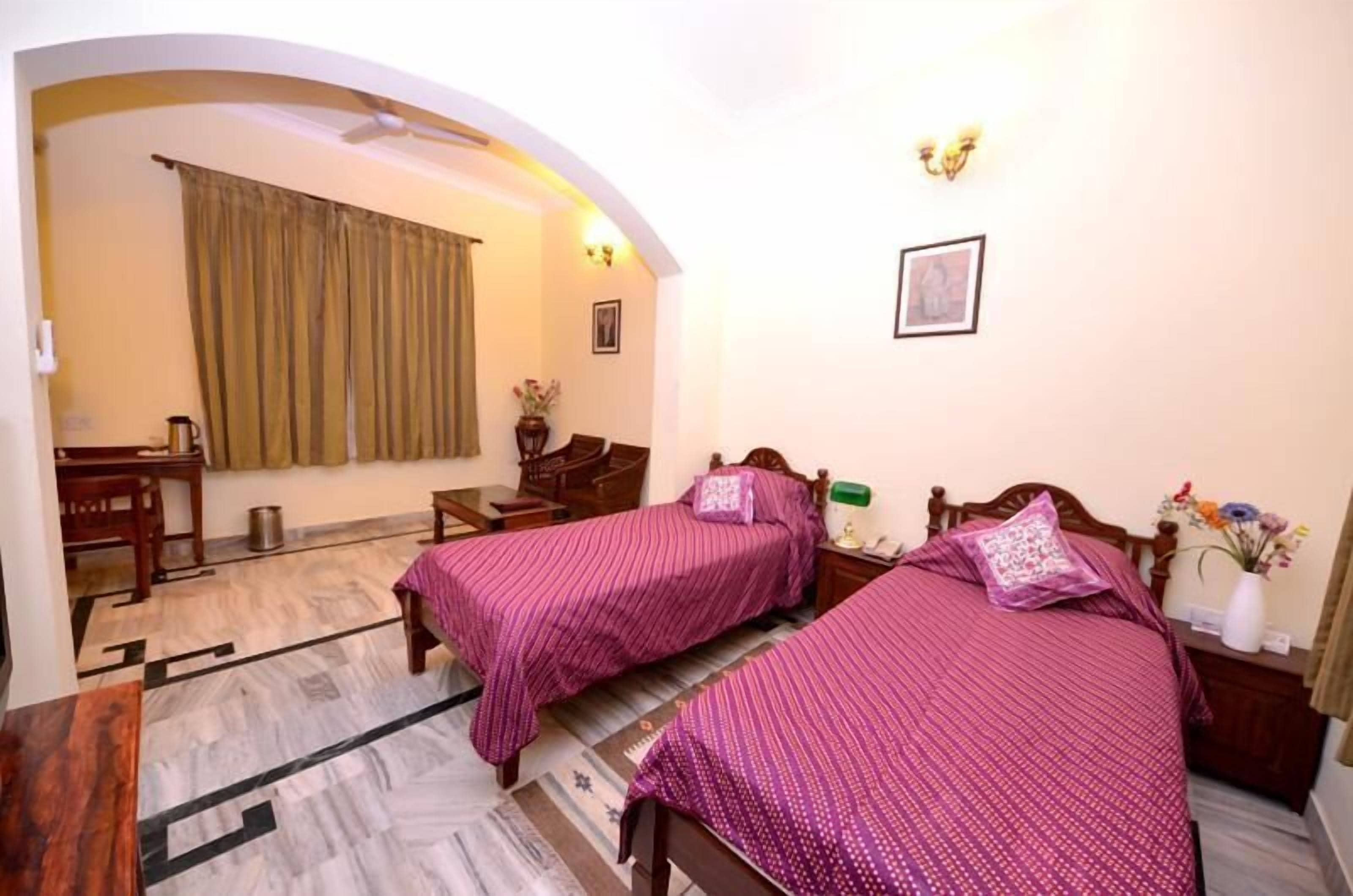 A room with two beds, a desk, a chair, a small table, and a window with curtains.