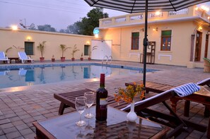 A poolside area with lounge chairs, a table with a bottle and glasses, and a building with a balcony in the background. - Koolwal Kothi Zinc Journey by The Fern (Nawalgarh)