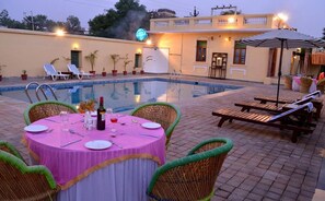 A poolside dining area with a table set for two, lounge chairs, and a pool. - Koolwal Kothi Zinc Journey by The Fern (Nawalgarh)