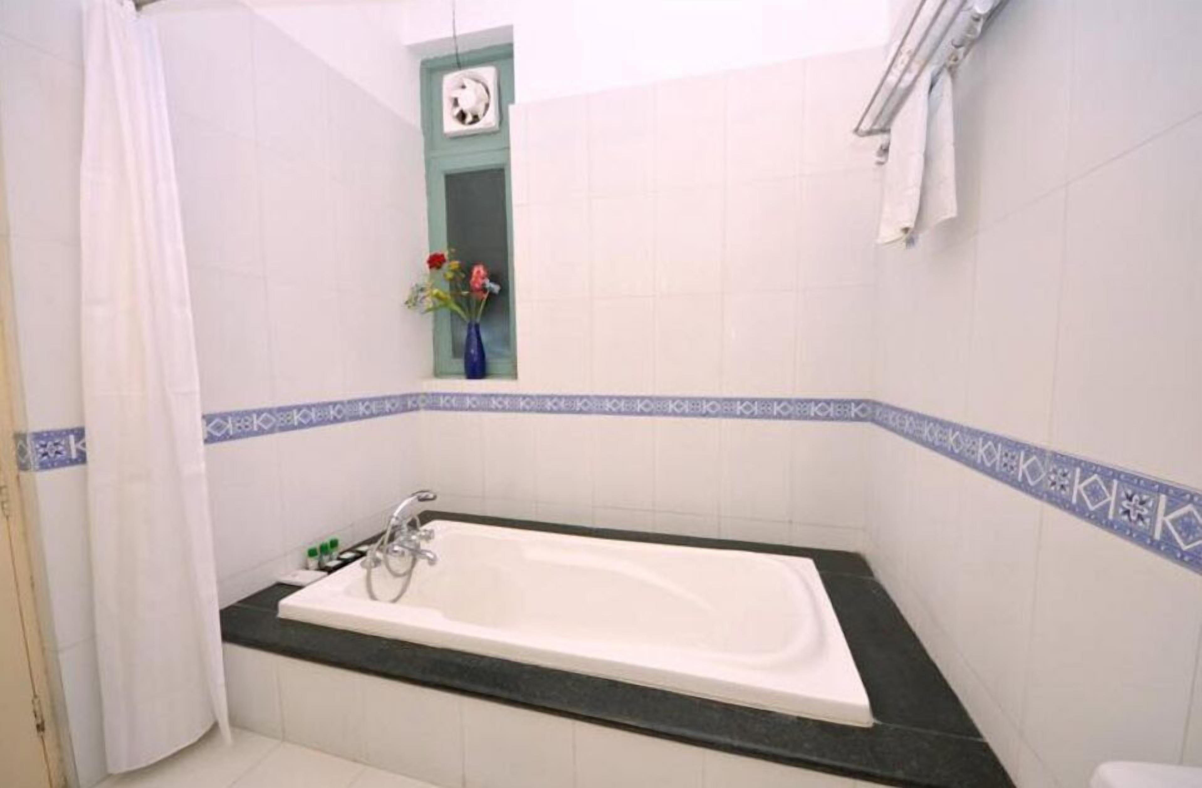 Een badkamer met een bad, douchegordijn, klein raam en bloemenvaas, en betegelde muren.