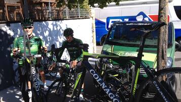 Un ciclista con una maglia verde seduto sulla bicicletta accanto a un veicolo parcheggiato con un logo ben visibile.