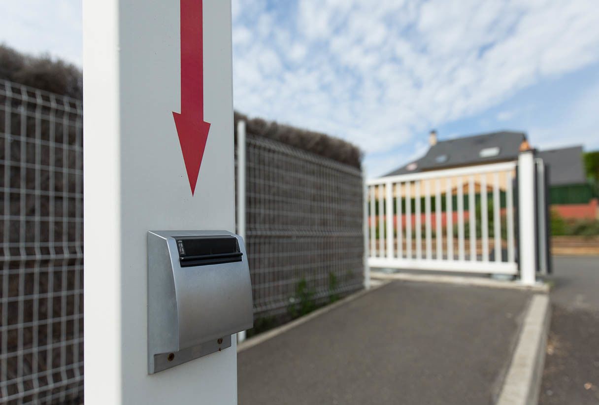A security gate with a red arrow pointing downwards, a metal box attached to the gate, and a building with a fence in the background.