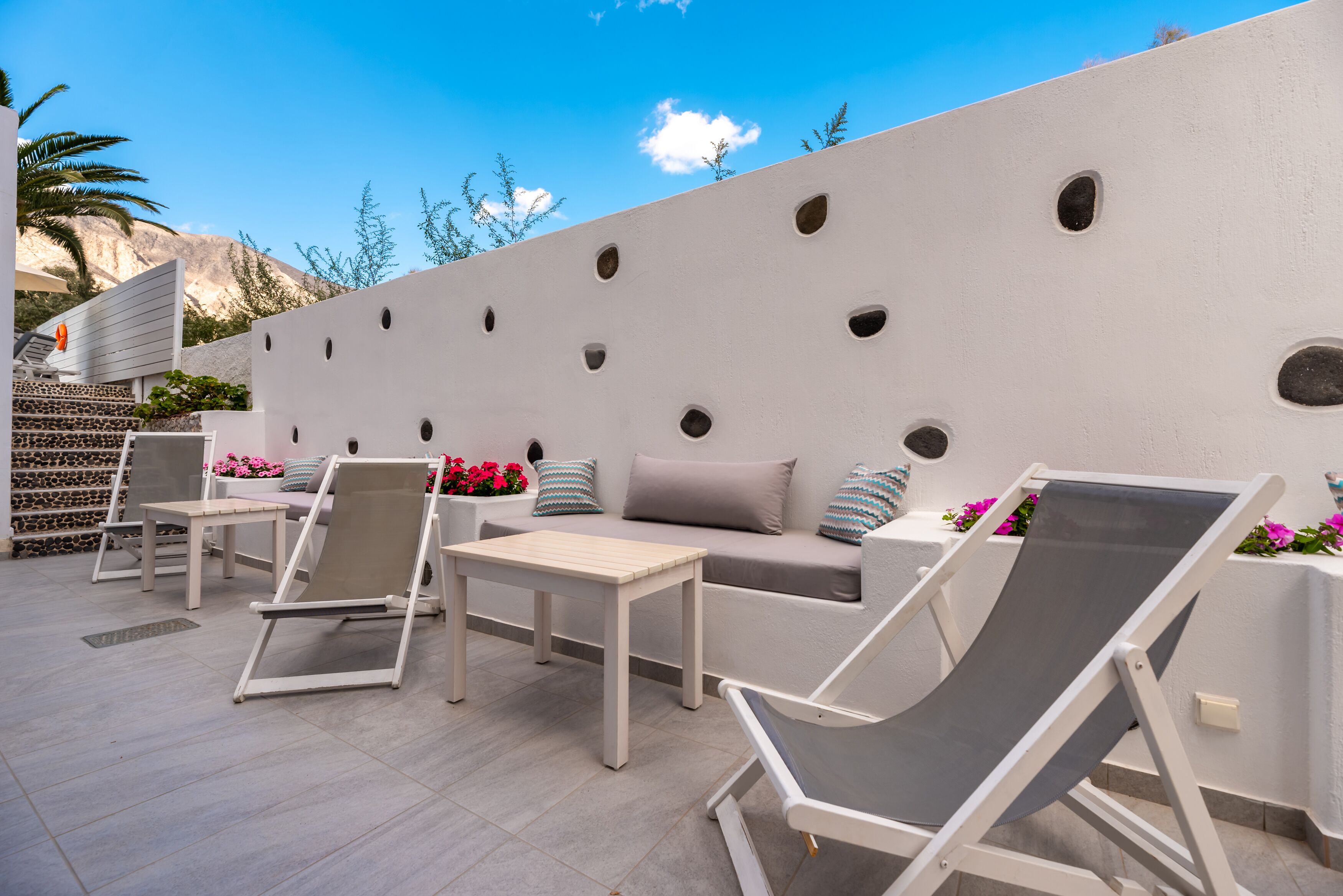 An outdoor seating area with a white wall, multiple lounge chairs, a small table, and decorative pillows.