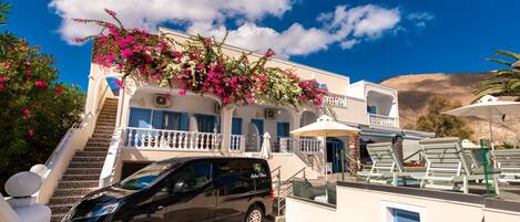 A white building with a balcony, a car parked in front, and a mountain in the background.