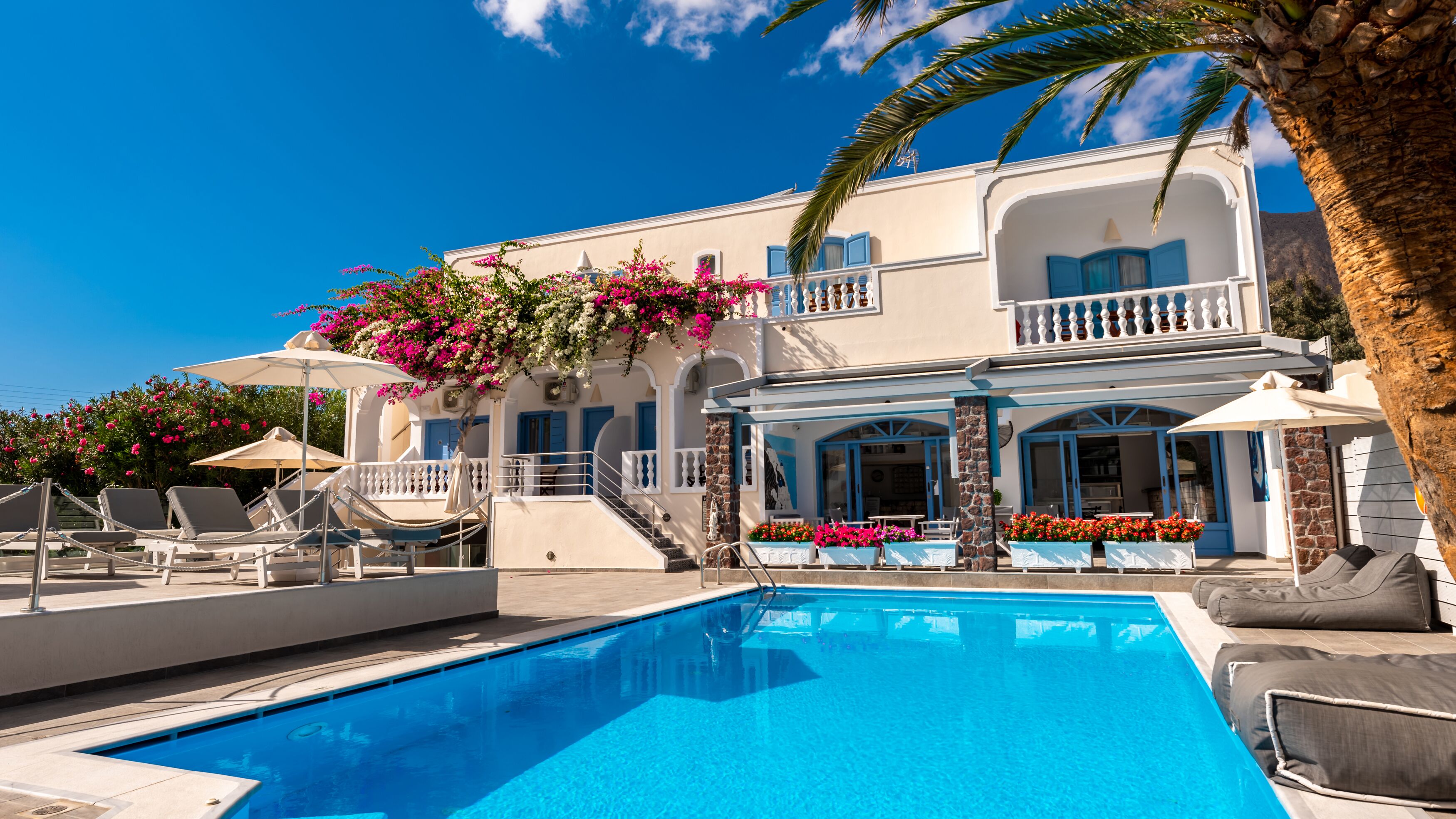 A poolside area with a clear blue pool, lounge chairs, and a building with blue shutters and white walls.