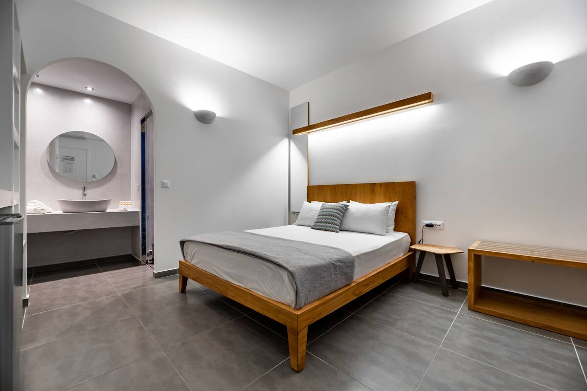 Un dormitori modern amb un llit de fusta, una taula de nit, una panca integrada i un bany amb un lavabo i un mirall.