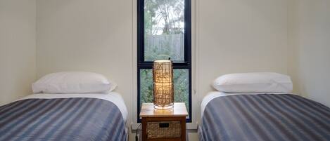Une chambre avec deux lits, une table de chevet, une lampe et une fenêtre donnant sur des arbres.