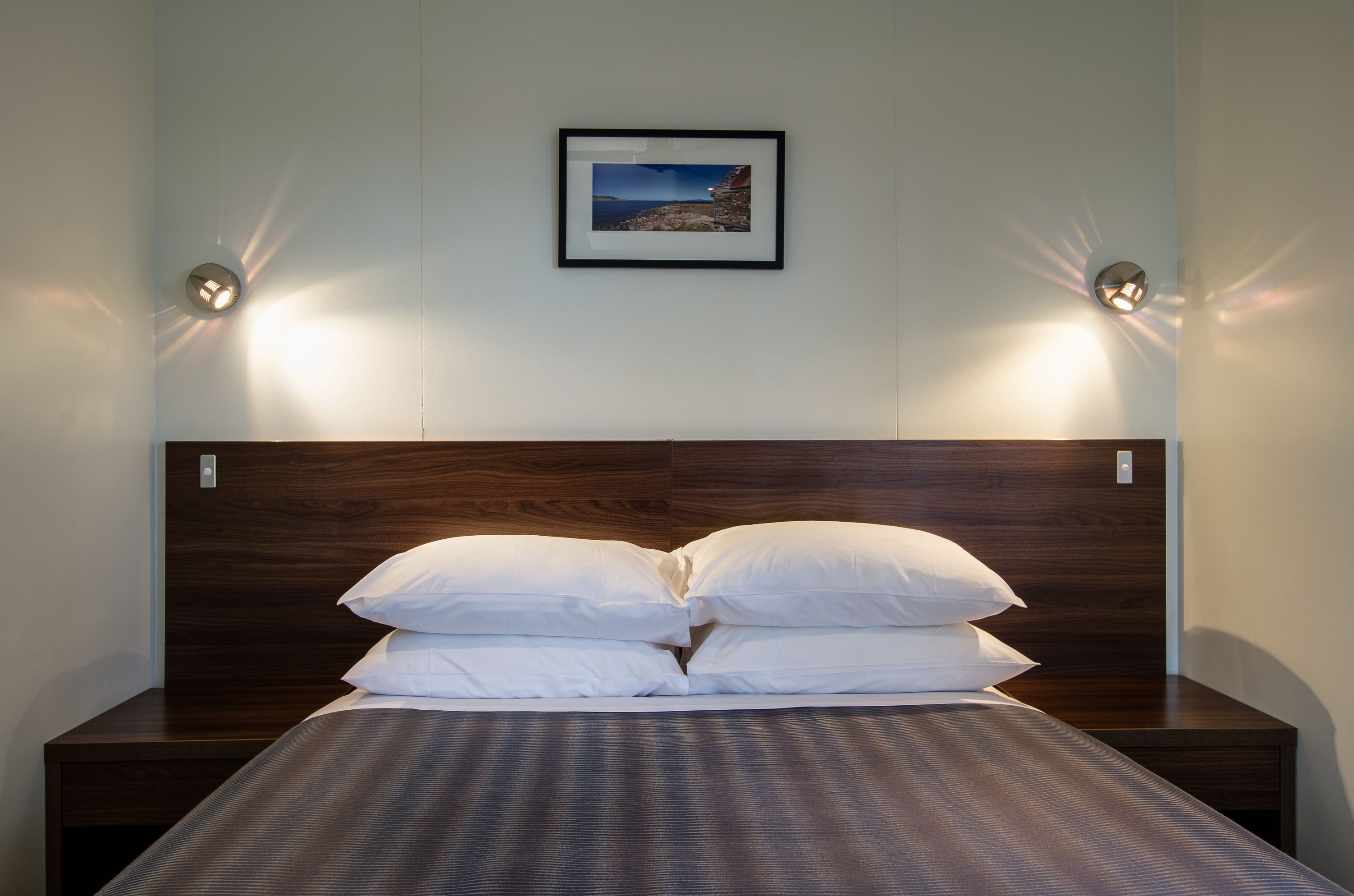 Una cama bien tendida con almohadas blancas y un cabecero oscuro, un cuadro en la pared y dos lámparas de pared a cada lado.