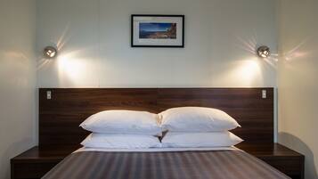Una cama bien tendida con almohadas blancas y un cabecero oscuro, un cuadro en la pared y dos lámparas de pared a cada lado.