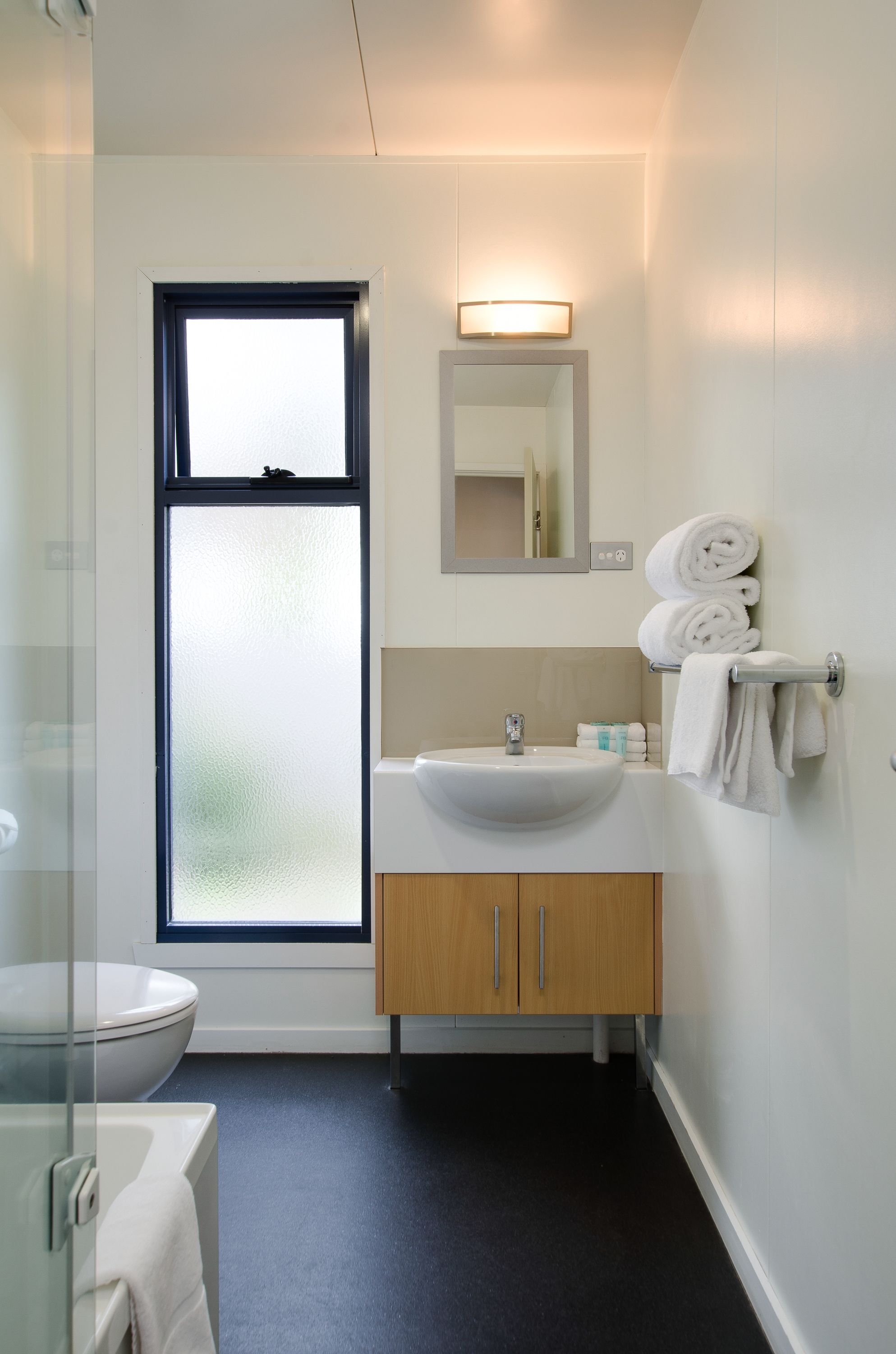 Un baño moderno con lavamanos blanco, armarios de madera, cabina de ducha con mampara de vidrio y una ventana con cristal satinado.