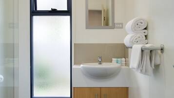 Un baño moderno con lavamanos blanco, armarios de madera, cabina de ducha con mampara de vidrio y una ventana con cristal satinado.