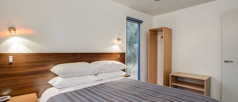 Een moderne hotelkamer met een houten hoofdbord, een groot bed met een gestreept dekbed, een nachtkastje met een thermometer, een garderobe en een plafondventilator.
