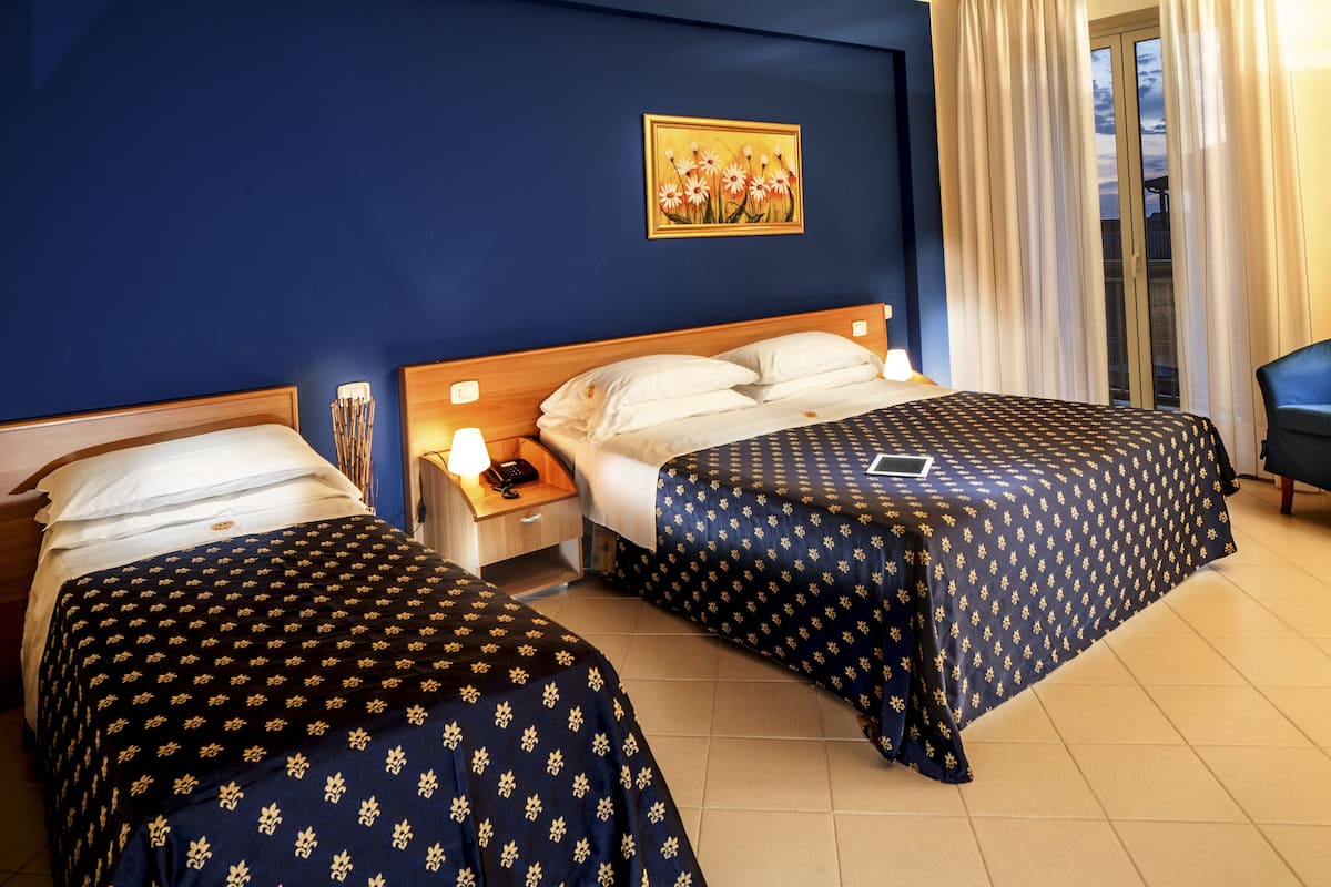 Habitación de hotel con dos camas, una mesita de noche con lámpara, un cuadro en la pared y vistas al exterior por la ventana.